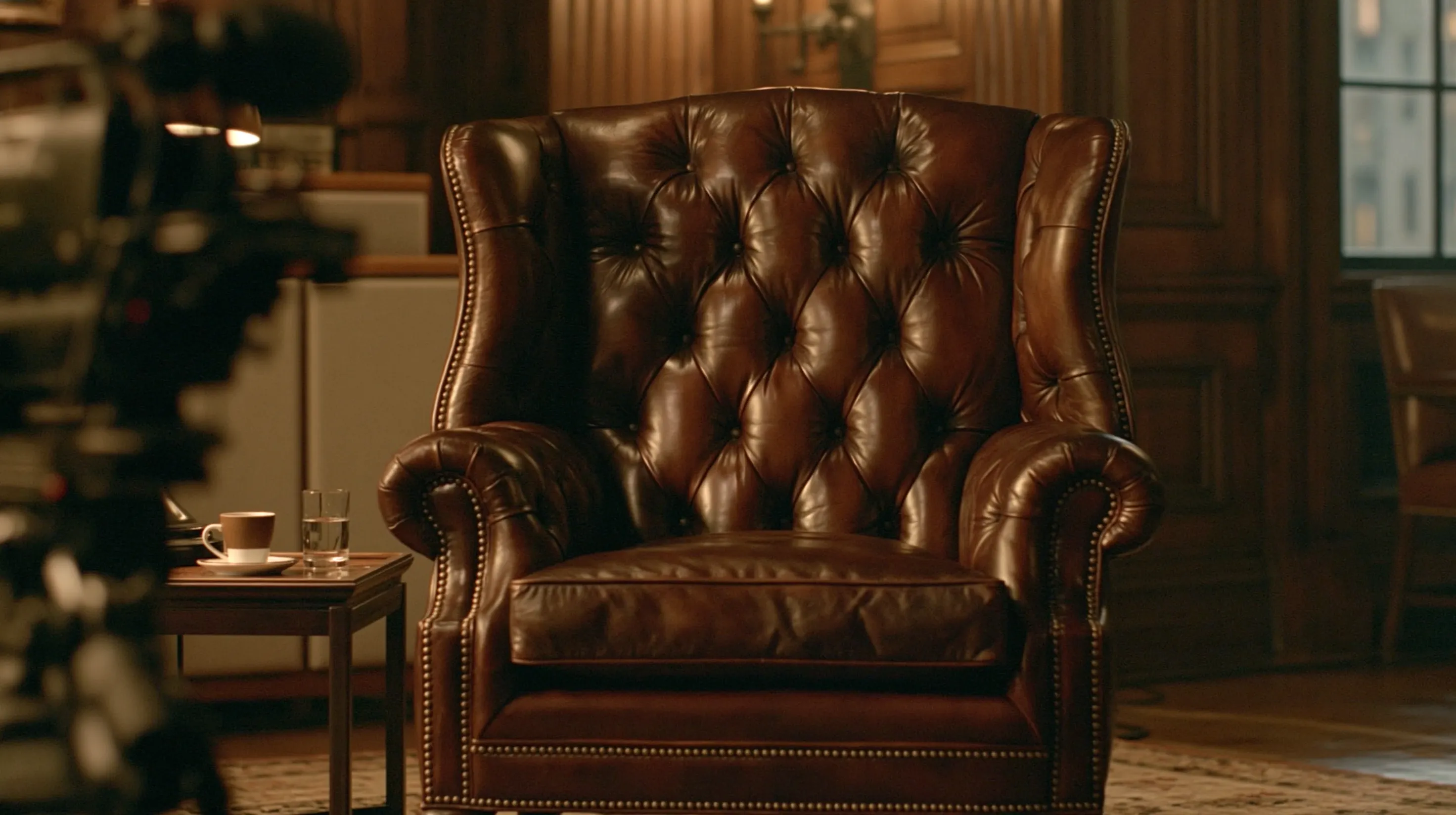 A leather armchair in the recording room under warm light, a cup of coffee and a glass of water on the side table, a camera just out of focus in the foreground.